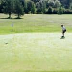 Trois jeunes golfeurs s'entraînent sur un green ensoleillé, concentrés sur leurs putts. L'herbe est bien entretenue et le drapeau est visible au centre du green. En arrière-plan, on aperçoit des arbres et un paysage verdoyant, créant une ambiance paisible propice à la pratique du golf. Cette image illustre l'importance de l'entraînement et de la concentration dans ce sport.