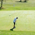 Un jeune golfeur en train de putter sur un green bien entretenu, sous un ciel ensoleillé. La scène montre la concentration du joueur alors qu'il vise le trou, avec un drapeau blanc en arrière-plan. L'environnement verdoyant du parcours de golf met en valeur l'activité de plein air, idéale pour les amateurs de golf et les passionnés de nature.