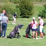 Un groupe de golfeurs observe un joueur sur le parcours, prêt à frapper sa balle. On aperçoit des enfants en tenue de golf, portant des polos roses, qui s'affairent autour de leurs chariots de golf. L'arrière-plan verdoyant met en valeur l'environnement naturel du terrain de golf. Des adultes, dont un homme en chapeau, regardent attentivement la scène, soulignant l'aspect social et convivial du golf. Cette image illustre parfaitement une
