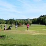Un groupe de golfeurs sur un parcours verdoyant par une journée ensoleillée. Deux jeunes joueuses, l'une en t-shirt rose et l'autre en tenue de golf, observent le jeu, tandis qu'un homme en tenue décontractée se prépare à frapper la balle. Des chariots de golf sont visibles à proximité, ajoutant à l'ambiance familiale du sport. En arrière-plan, un paysage naturel avec des arbres et des collines complète la scène tranquille d'une