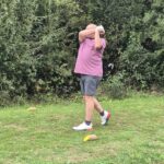 Un homme en polo rose s'apprête à frapper une balle de golf sur un terrain verdoyant. Il se tient sur une pelouse bien entretenue, entouré de buissons. Des cônes de signalisation orange et jaune sont disposés au sol, probablement pour un exercice de golf. L'image illustre une activité sportive en plein air, mettant en avant la concentration et la technique dans le golf.