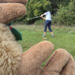 Image d'un personnage en peluche tenant une batte de baseball, avec une personne en arrière-plan qui frappe une balle sur un terrain verdoyant. La scène se déroule en plein air, entourée d'arbres et de verdure, illustrant une activité sportive conviviale. Cette image évoque le plaisir du baseball et les moments de loisirs en famille ou entre amis.
