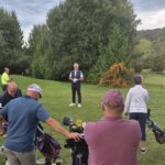 Groupe de golfeurs assistant à une séance d'entraînement sur un parcours verdoyant. Un instructeur, vêtu d'une tenue de golf, donne des conseils à un public attentif. Les participants, de différents âges et styles vestimentaires, écoutent attentivement tout en tenant leurs sacs de golf. En arrière-plan, une golfeuse s'exerce au putting près du drapeau. L'image illustre l'esprit communautaire et l'apprentissage dans le golf.