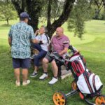 Groupe d'amis se détendant sur un parcours de golf verdoyant, profitant d'une pause après une partie. L'un d'eux est en train de manger, tandis que les autres échangent des anecdotes. Un chariot de golf est visible à côté d'eux, illustrant l'ambiance conviviale et décontractée d'une journée de golf. En arrière-plan, d'autres golfeurs sont engagés dans leur jeu, ajoutant à l'animation du lieu.