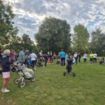 Groupe de golfeurs rassemblés sur un parcours, écoutant un instructeur. Plusieurs participants sont équipés de chariots de golf, tandis que d'autres se tiennent debout, attentifs. L'environnement est verdoyant, avec des arbres en arrière-plan et un ciel partiellement nuageux. Cette scène illustre une activité de golf en plein air, favorisant le sport et la camaraderie.