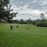Groupe de golfeurs sur un parcours verdoyant, marchant avec des sacs de golf. Certains utilisent des parapluies pour se protéger d'une légère pluie, tandis que le ciel est nuageux. L'environnement naturel est entouré d'arbres et de buissons, créant une ambiance paisible pour la pratique du golf. Cette image illustre une journée de golf en plein air, idéale pour les amateurs de ce sport.