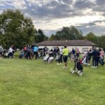 Groupe de golfeurs rassemblés sur un terrain de golf, avec des chariots de golf et des sacs visibles. L'environnement est verdoyant, avec des arbres et un bâtiment en arrière-plan. L'atmosphère est conviviale et dynamique, indiquant une compétition ou un événement de golf en cours. Le ciel est partiellement nuageux, créant une belle lumière pour une journée de golf.