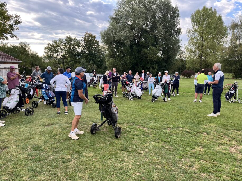 Groupe de golfeurs sur un parcours, rassemblés pour un briefing avant le départ. Plusieurs participants, hommes et femmes, sont équipés de sacs de golf sur des chariots. L'ambiance est conviviale et dynamique, avec des arbres en arrière-plan et un ciel nuageux. Cet événement met en avant la passion du golf et le partage entre amateurs du sport.