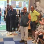 Un groupe de personnes assiste à une réunion conviviale dans un espace de loisirs, où un homme prend la parole pour partager des informations. Les participants, de différents âges, sont attentifs et engagés, certains assis, d'autres debout. L'arrière-plan présente des affiches et des équipements, créant une ambiance dynamique. Cette scène illustre un moment de partage et d'interaction au sein d'une communauté.