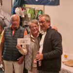 Événement convivial dans un club de golf où des participants célèbrent leurs succès. Trois personnes, souriantes, posent ensemble avec un prix en main, tandis qu'un homme assis au fond semble apprécier l'ambiance. En arrière-plan, une image d'un golfeur célèbre et des accessoires de golf sont exposés. L'atmosphère est chaleureuse et festive, reflétant l'esprit communautaire du club.