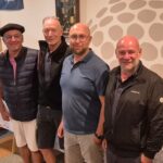 Quatre hommes se tiennent côte à côte dans un espace de rassemblement, probablement lors d'un événement de golf. Ils affichent des sourires et portent des vêtements décontractés adaptés à une activité sportive. En arrière-plan, on aperçoit des accessoires de golf et des bouteilles de vin sur une table, suggérant une ambiance conviviale. L'image reflète un moment de camaraderie et de célébration, typique des événements de golf où les participants se réunissent après une compétition