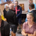 Groupe de personnes rassemblées dans un espace intérieur, discutant et interagissant. On peut voir des hommes et des femmes de différents âges, vêtus de tenues décontractées. L'environnement est chaleureux, avec des sièges en bois et des éléments décoratifs sur les murs. Cette image illustre un moment convivial, propice aux échanges et à la socialisation.