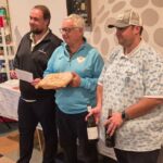 Photo d'une remise de prix lors d'un événement convivial, avec des participants rassemblés autour d'une table. Trois hommes se tiennent au premier plan, l'un d'eux tenant un prix, tandis que d'autres participants sont assis en arrière-plan, visiblement attentifs. Plusieurs bouteilles de vin sont disposées sur la table, suggérant une célébration ou un concours lié à la gastronomie ou aux loisirs. L'ambiance est détendue et amicale, typique des