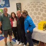 Cérémonie de remise de prix au club de golf, avec plusieurs participants rassemblés autour d'une table garnie de bouteilles de vin et de récompenses. En arrière-plan, des décorations liées au golf, dont un poster de la Ryder Cup 2018. L'ambiance est conviviale, avec des membres du club engagés dans des discussions, soulignant l'esprit communautaire du golf.