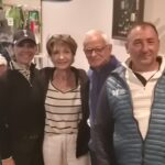 Quatre personnes posent ensemble dans un cadre convivial, probablement lors d'un événement social. En arrière-plan, plusieurs participants sont assis, créant une atmosphère animée. La tenue des personnes suggère un contexte sportif, possiblement lié au golf, avec des vêtements adaptés à l'activité. L'image évoque la camaraderie et le plaisir partagé entre amis ou membres d'un club.