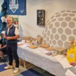 Événement de golf avec des participants réunis dans une salle, écoutant un discours. À l'arrière-plan, une affiche de la Ryder Cup 2018. Des tables sont garnies de prix et de produits divers. L'ambiance est conviviale et dynamique, illustrant l'esprit communautaire autour du golf.