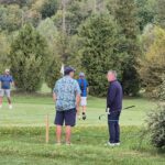 Un groupe de golfeurs se rassemble sur un parcours verdoyant, entouré d'arbres majestueux. Deux hommes discutent près d'un green, tandis que d'autres joueurs se préparent à frapper leur balle. L'atmosphère est détendue et conviviale, idéale pour une journée de golf en pleine nature. Des éléments tels que des drapeaux de parcours et des équipements de golf ajoutent à l'ambiance sportive de cette scène.