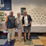Trois participants souriants posent ensemble dans un club de golf, célébrant un événement lié à la Ryder Cup 2018. L'arrière-plan présente une décoration de golf, incluant un drapeau de la Ryder Cup et des articles de golf en vente. L'un des participants tient une bouteille de vin, tandis qu'un autre montre un document, probablement un prix ou une reconnaissance. Cette image évoque l'esprit convivial et la camaraderie des passionnés de golf lors d'une compétition