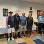 Quatre hommes se tiennent côte à côte dans un cadre convivial, probablement lors d'un événement ou d'une remise de prix. Ils portent des tenues décontractées, adaptées à une journée sportive. En arrière-plan, on peut apercevoir une table décorée et des affiches sur les murs, suggérant une ambiance festive. Des participants sont visibles à une table à droite, ajoutant à l'atmosphère dynamique de l'événement. Cette image illustre une rencontre sociale autour du