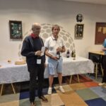 Cérémonie de remise de prix dans un club de golf, mettant en avant deux hommes souriants tenant des bouteilles de vin. En arrière-plan, une table décorée avec des récompenses et des décorations. L'ambiance est conviviale, illustrant la célébration des performances des participants. Les murs sont ornés d'objets liés au golf, soulignant l'esprit sportif de l'événement. Cette image capture l'essence d'une journée mémorable dédiée au golf