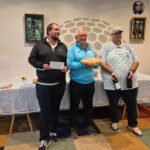 Cérémonie de remise de prix avec trois participants souriants. Le premier homme tient une pancarte, le deuxième, portant un pull bleu, présente un gâteau, tandis que le troisième, en chemise à motifs, tient deux bouteilles de vin. En arrière-plan, une table est décorée avec une nappe et des objets, suggérant une ambiance conviviale. Événement organisé par le Crédit Mutuel, mettant en avant l'esprit de camaraderie et de célébration.
