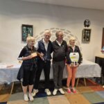 Groupe de participants à un événement de golf, posant ensemble après une remise de prix. Ils tiennent des récompenses et affichent des sourires, symbolisant l'esprit sportif et l'amitié. En arrière-plan, des éléments décoratifs et des tables avec des produits sponsorisés, créant une ambiance conviviale. Cette image illustre l'importance des compétitions de golf et des moments de partage entre passionnés.