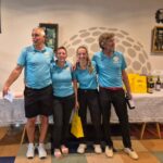 Quatre participants se tiennent côte à côte dans un espace convivial, souriant et vêtus de polos bleus. Ils semblent célébrer un événement lié au golf, entourés de bouteilles de vin et de cadeaux sur une table. L'ambiance est festive, suggérant une remise de prix ou une compétition. Le décor présente des éléments évoquant le golf, renforçant le thème de l'événement.