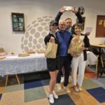 Groupe de personnes souriantes lors d'une remise de prix dans un club de golf. Une femme en tenue de golf tient un sac, tandis que d'autres membres du groupe tiennent des cadeaux. En arrière-plan, une table avec des articles et un tableau d'affichage. Ambiance conviviale et festive, illustrant un événement communautaire de golf.