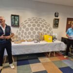 Deux hommes sont présents dans une salle de réunion, l'un s'exprimant tandis que l'autre écoute attentivement. Ils se trouvent devant une table décorée, chargée de divers prix et cadeaux, incluant des bouteilles de vin et des sacs colorés. L'environnement est convivial, avec des éléments décoratifs liés au golf sur les murs. Cette scène évoque une remise de prix ou un événement social autour du golf, soulignant l'importance de la camaraderie et de la célébr