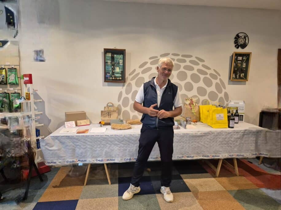 Homme debout dans une salle de réception, souriant, vêtu d'un gilet et d'un polo. À l'arrière-plan, une table décorée avec divers articles, y compris des bouteilles de vin, des sacs jaunes et des objets de décoration. Mur orné d'images et de motifs de golf, créant une ambiance conviviale. Cette image illustre un événement social ou une remise de prix dans un cadre sportif.