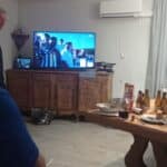 Groupe d'amis réunis dans un salon chaleureux, regardant un événement à la télévision. L'écran affiche des spectateurs avec des drapeaux, tandis que des bouteilles et des verres sont disposés sur la table en bois. L'ambiance conviviale est accentuée par des mets et des boissons, suggérant un moment de partage et de convivialité.