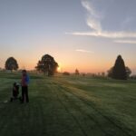 Un golfeur se tient sur un parcours de golf au lever du soleil, entouré de verdure et d'arbres majestueux. La brume matinale enveloppe le terrain, créant une atmosphère paisible. Un autre joueur s'éloigne, ses clubs de golf à la main. Cette image évoque la beauté d'une matinée de golf, idéale pour les amateurs de ce sport. Les couleurs chaudes du ciel ajoutent une touche magique à ce moment.