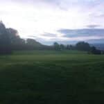 Vue panoramique d'un green de golf au lever du soleil, entouré d'arbres verdoyants. Le drapeau blanc indique le trou, tandis que le ciel montre des nuances de bleu et de gris. Ce cadre paisible est idéal pour les amateurs de golf et les passionnés de nature. Parfait pour une journée de jeu ou une promenade tranquille sur le parcours.