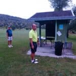 Deux golfeurs se tiennent près d'un abri sur un parcours de golf, entourés d'un paysage verdoyant et de collines en arrière-plan. L'un des golfeurs porte un haut à manches courtes de couleur vive, tandis que l'autre est habillé de manière décontractée avec un équipement adapté au golf. L'abri est équipé d'une table et d'une poubelle, et des panneaux d'information sont visibles. Cette scène illustre un moment de détente