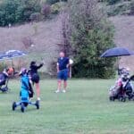 Groupe de golfeurs sur un parcours, avec une femme en train de frapper une balle et deux autres personnes observant. Des chariots de golf sont visibles, ainsi qu'un parasol pour se protéger du soleil. L'environnement est verdoyant, avec des collines en arrière-plan, créant une ambiance relaxante propice au golf.
