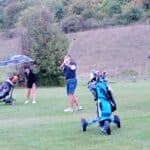 Un groupe de golfeurs sur un parcours verdoyant, malgré une météo nuageuse. Un homme s'apprête à frapper la balle avec un club de golf, tandis qu'une femme observe sous un parapluie. Deux autres golfeurs, l'un tenant un parapluie et l'autre poussant une voiturette de golf, se déplacent sur le terrain. Les équipements de golf, dont des sacs et des clubs, sont visibles, témoignant de l'ambiance conviviale