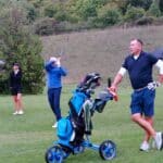 Un groupe de golfeurs sur un parcours verdoyant, avec des arbres en arrière-plan. Un homme en polo noir se tient près d'un chariot de golf, tandis qu'un autre homme s'apprête à frapper une balle. Deux femmes, l'une sous un parapluie et l'autre en tenue de golf, observent la scène. L'image capture l'ambiance décontractée d'une journée de golf, mettant en avant l'équipement de golf et le paysage naturel.