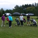 Groupe de golfeurs sur un parcours verdoyant, utilisant des parapluies pour se protéger de la pluie. Ils sont accompagnés de chariots de golf, prêts à jouer malgré les conditions météorologiques. L'image illustre une journée de golf en plein air, mettant en avant l'esprit d'équipe et la passion pour ce sport.