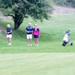 Quatre golfeurs se tiennent sur un parcours verdoyant, entourés d'arbres. Deux hommes et deux femmes, vêtus de vêtements de golf adaptés, semblent discuter et profiter d'une journée en plein air. Un chariot de golf est garé à proximité, indiquant qu'ils sont en train de jouer ou de se préparer à jouer. L'image capture l'ambiance conviviale et relaxante d'une partie de golf, illustrant le plaisir du jeu en groupe.