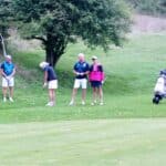 Quatre golfeurs se tiennent sur un parcours verdoyant, entourés de nature. Un joueur s'apprête à frapper sa balle, tandis que les autres observateurs attendent avec intérêt. L'un d'eux, vêtu d'un polo bleu, tient une balle dans sa main, et une golfeuse en vêtement rose sourit en attendant son tour. À côté d'eux, un chariot de golf est prêt pour la prochaine partie. Cette scène illustre l