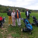 Quatre golfeurs se tiennent sur un parcours verdoyant, entourés de collines boisées. Ils sont équipés de sacs de golf, prêts à jouer. L'un porte un chapeau bleu, tandis qu'une femme au centre tient un club de golf. Les autres hommes portent des tenues variées, allant du polo à un gilet. L'atmosphère est conviviale et décontractée, idéale pour une journée de golf entre amis.