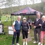Quatre golfeurs posent ensemble sur un parcours de golf, avec un stand de la marque 24MX en arrière-plan. On aperçoit des chariots de golf et d'autres joueurs sur le green, entourés de verdure. Le groupe sourit et semble profiter d'une journée ensoleillée. Le panneau indiquant le restaurant "Le Cottage" ajoute un élément local à cette scène conviviale de golf.