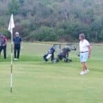 Trois golfeurs sur le green d'un parcours de golf, avec un drapeau blanc indiquant le trou. Deux joueurs marchent vers le trou, tandis qu'un troisième se dirige vers eux. Des sacs de golf sont visibles en arrière-plan. Le paysage environnant est verdoyant, avec des arbres en arrière-plan. Cette scène illustre l'ambiance conviviale et relaxante d'une journée de golf.