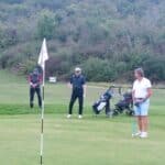 Trois golfeurs sur un green, se préparant à jouer. Un joueur est en train de putter près du drapeau, tandis que les autres observent. Des sacs de golf sont visibles à proximité, sur un parcours verdoyant entouré de végétation. Cette scène illustre une journée de golf agréable en plein air.