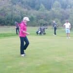 Un groupe de golfeurs sur un parcours verdoyant, se concentrant sur leur jeu. L'image montre quatre personnes, dont une femme en veste rose, en train de se préparer à putter. En arrière-plan, deux autres golfeurs s'entraînent avec leurs clubs, tandis qu'un chariot de golf est stationné à proximité. L'atmosphère est détendue, idéale pour une journée de golf en plein air. Cette scène illustre l'esprit convivial et sportif du golf