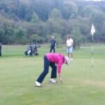 Un groupe de golfeurs sur un parcours verdoyant, mettant en avant une femme en train de récupérer sa balle sur le green. À l'arrière-plan, d'autres golfeurs se préparent à jouer, tandis que des sacs de golf sont visibles. L'environnement est paisible, avec des arbres en arrière-plan, illustrant une journée agréable de golf. Cette image capture l'esprit convivial de ce sport en plein air.