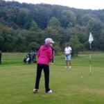Quatre golfeurs sur un parcours verdoyant, pratiquant le putting. Une femme en veste rose se prépare à frapper la balle, tandis que deux autres golfeurs observent sur le green. Un joueur en arrière-plan s'apprête à faire un coup avec un club. Des sacs de golf sont visibles, et un drapeau blanc indique le trou à proximité. L'environnement est calme, avec des arbres en arrière-plan, créant une atmosphère agréable pour le golf.