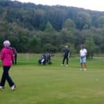 Un groupe de golfeurs sur le green d'un parcours de golf, avec une femme en veste rose marchant vers le trou. Deux hommes, l'un en polo blanc et l'autre en veste noire, observent attentivement la balle près du drapeau. Des sacs de golf sont visibles en arrière-plan, avec un paysage boisé en toile de fond, évoquant une journée de golf relaxante en plein air. Cette image illustre l'esprit convivial et compétitif du golf.