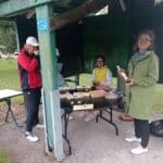Groupe de personnes réunies sous un abri de jardin, discutant et partageant des produits locaux. Sur la table, des boîtes en bois contenant des plantes succulentes et d'autres articles. Une femme souriante tient une bouteille, tandis qu'une jeune fille s'assoit à côté d'elle. L'ambiance est conviviale et décontractée, typique des marchés ou événements communautaires en plein air. L'environnement verdoyant et le mobilier simple ajoutent à l'attr