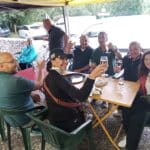 Groupe d'amis réunis sous un abri, levant leurs verres pour un toast. L'ambiance est festive, avec des rires et des sourires autour d'une table colorée. Les participants, vêtus de tenues décontractées, semblent profiter d'un moment convivial après une activité en plein air, probablement un barbecue ou un pique-nique. En arrière-plan, des voitures sont garées, indiquant un cadre paisible en nature. Cette image évoque des souvenirs
